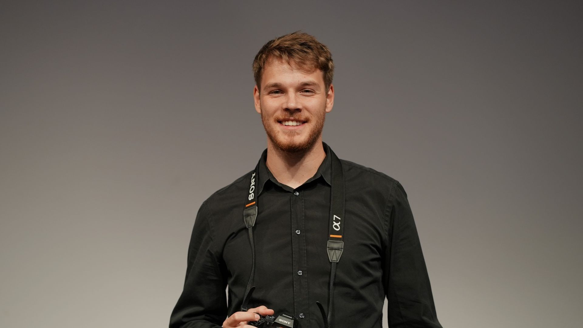 Man smiling, wearing a black shirt and holding a camera with a Sony strap around his neck.