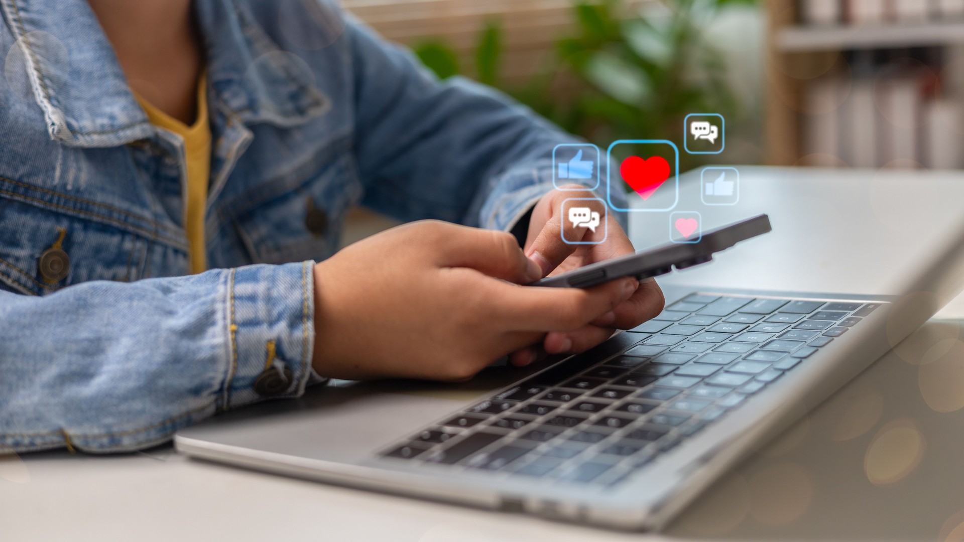 Using a smartphone while a laptop is open, with social media icons floating, such as likes, hearts, and comments, representing online interaction and digital lifestyle.